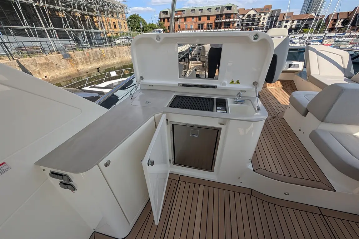 Outdoor boat galley with a grill, sink, and fridge, wooden decking, and marina view under a clear blue sky.
